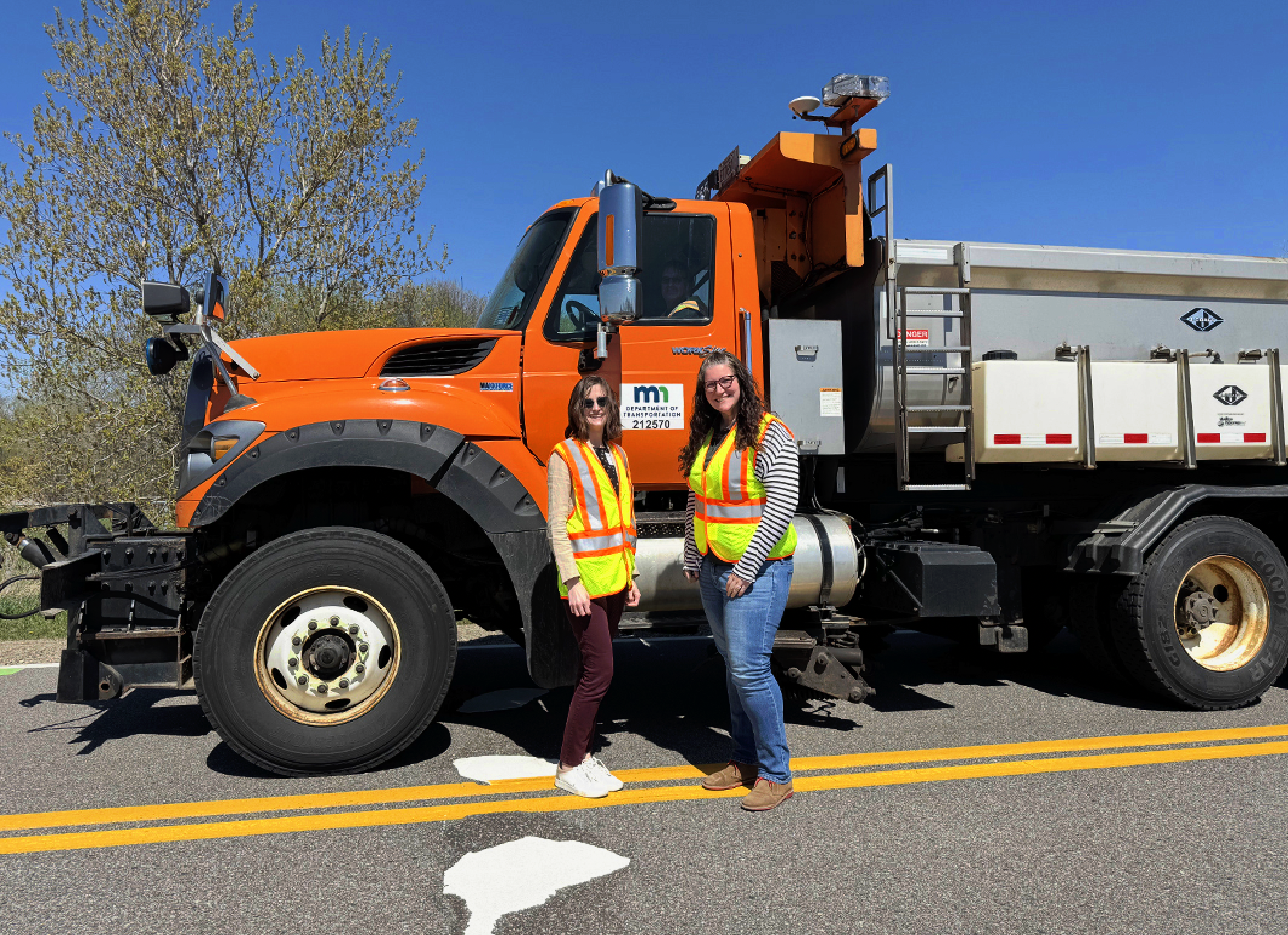 Two women in safety vests stand near a snowplow truck with the following text on the door: M.n. Department of Transportation 2 1 2 5 7 0